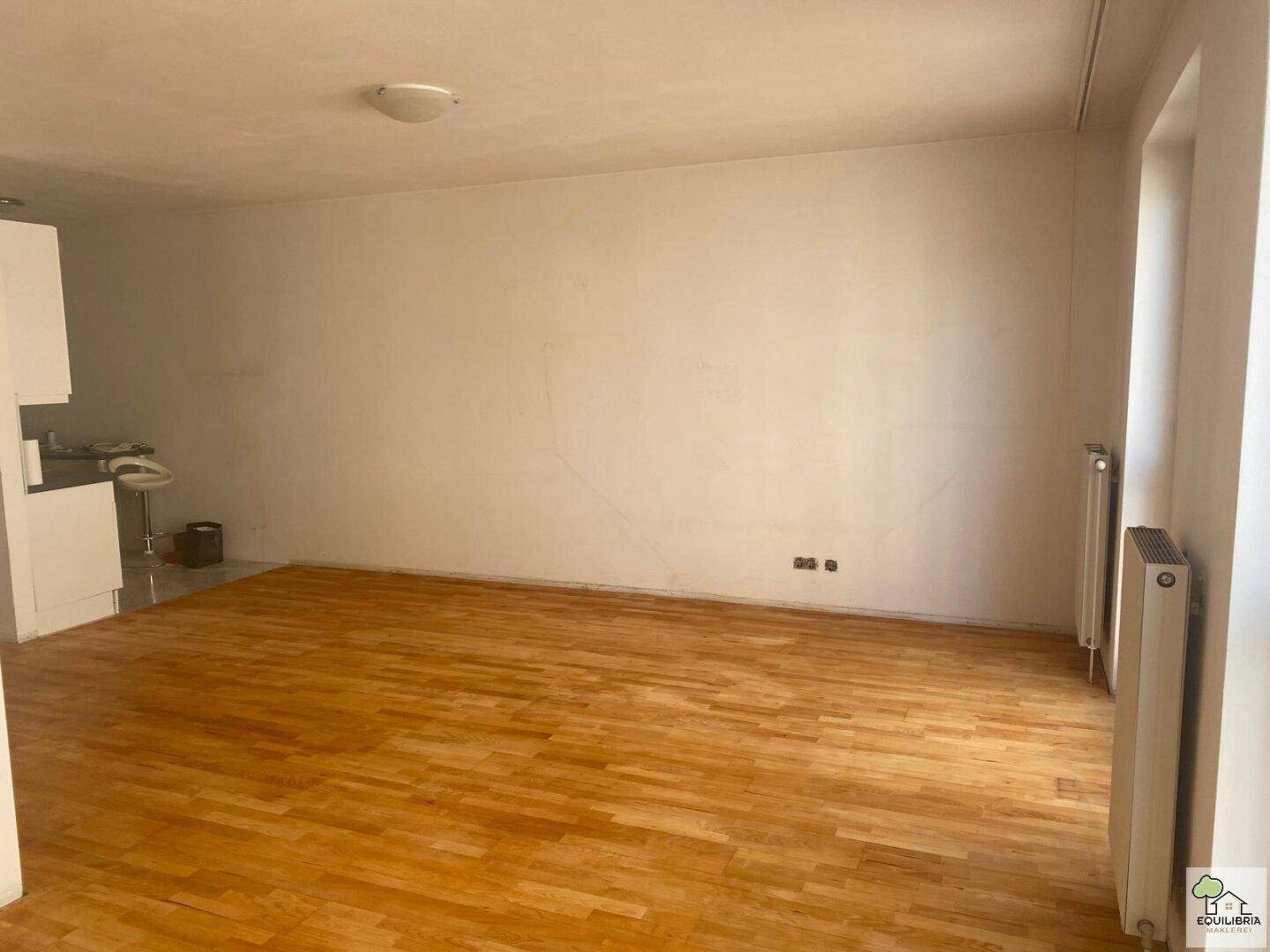 Offener Wohnbereich mit Parkettboden und Heizkörper, im Hintergrund ist ein Teil der Küche sichtbar.