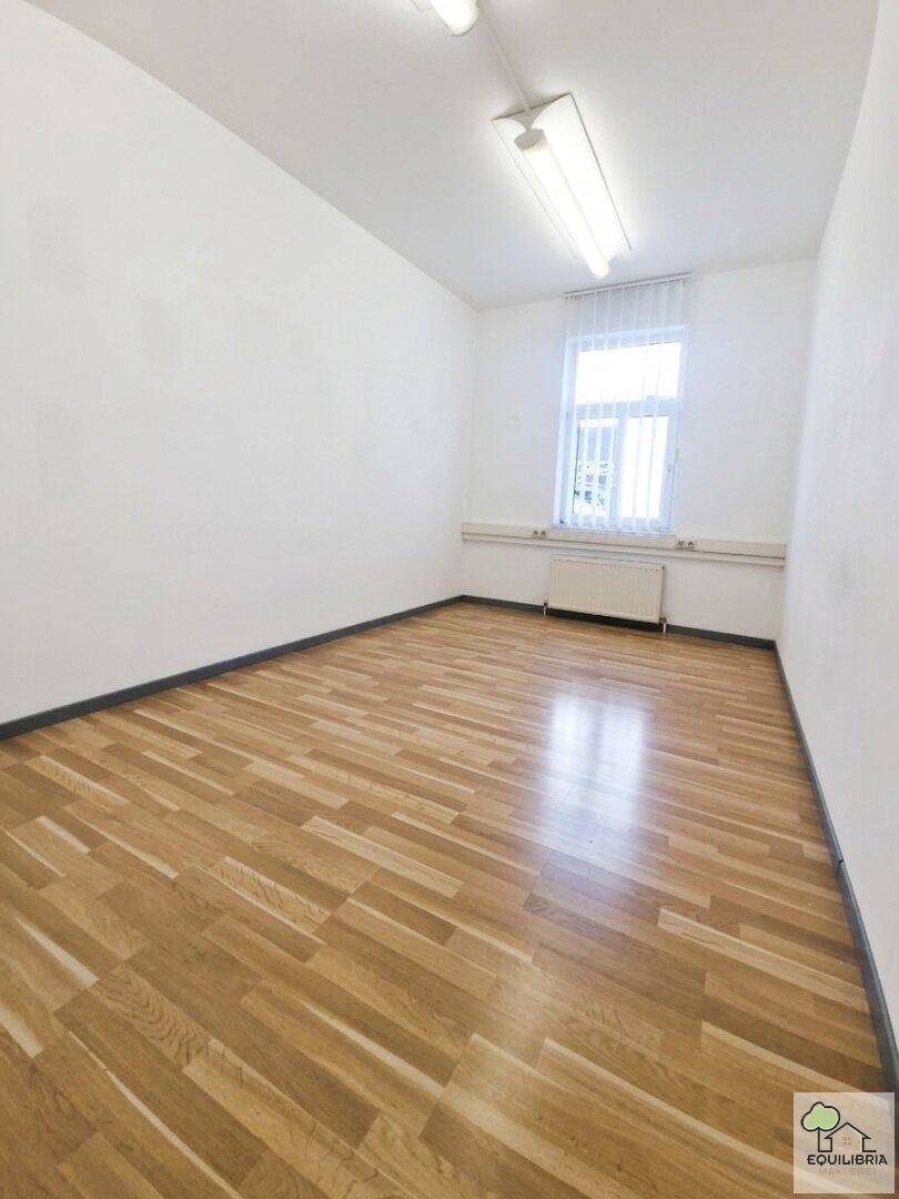 Leerer Raum mit hellem Holzparkett, weißen Wänden und einem Fenster mit Jalousien.