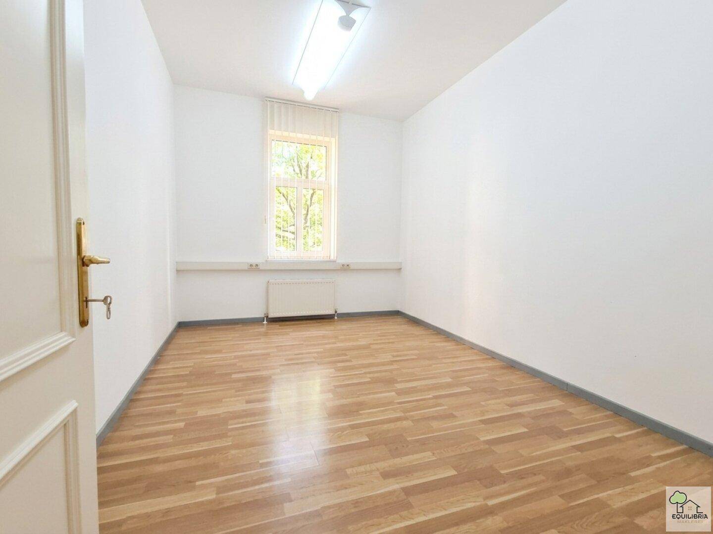 Kleinerer Büroraum mit hellem Holzfußboden, weiß gestrichenen Wänden und einem Fenster.