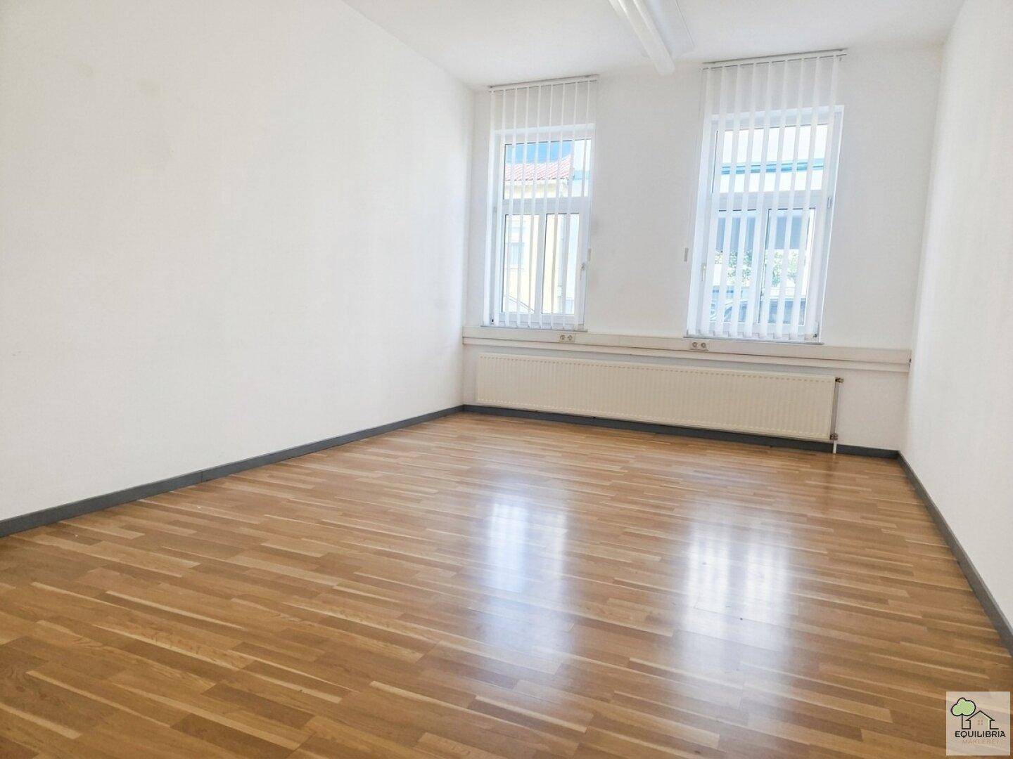 Geräumiger Büroraum mit hellem Holzfußboden, weißen Wänden und zwei Fenstern mit Jalousien.