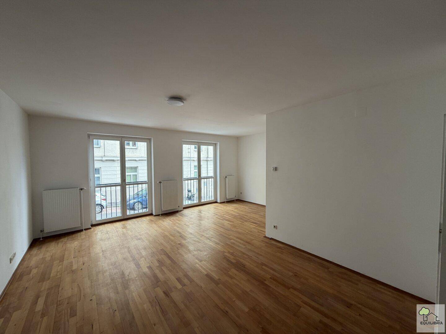 Geräumiges Wohnzimmer mit Holzboden, großen Fenstern und Blick auf die Straße.