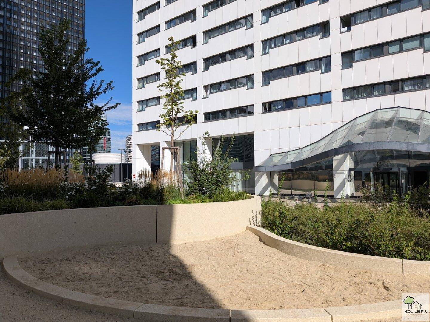 Sandkasten und begrünte Außenanlage vor einem modernen Gebäude mit großen Fenstern.