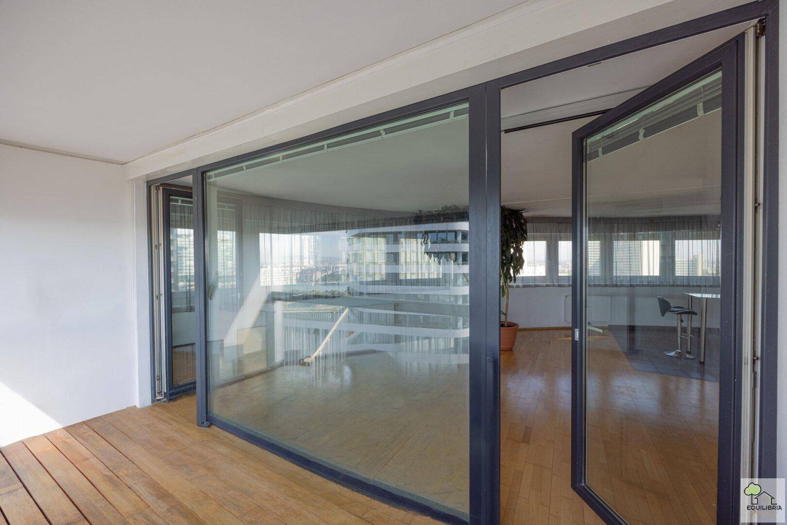 Geräumiger Wohnbereich mit Holzboden und großen Glasfronten zum Balkon.