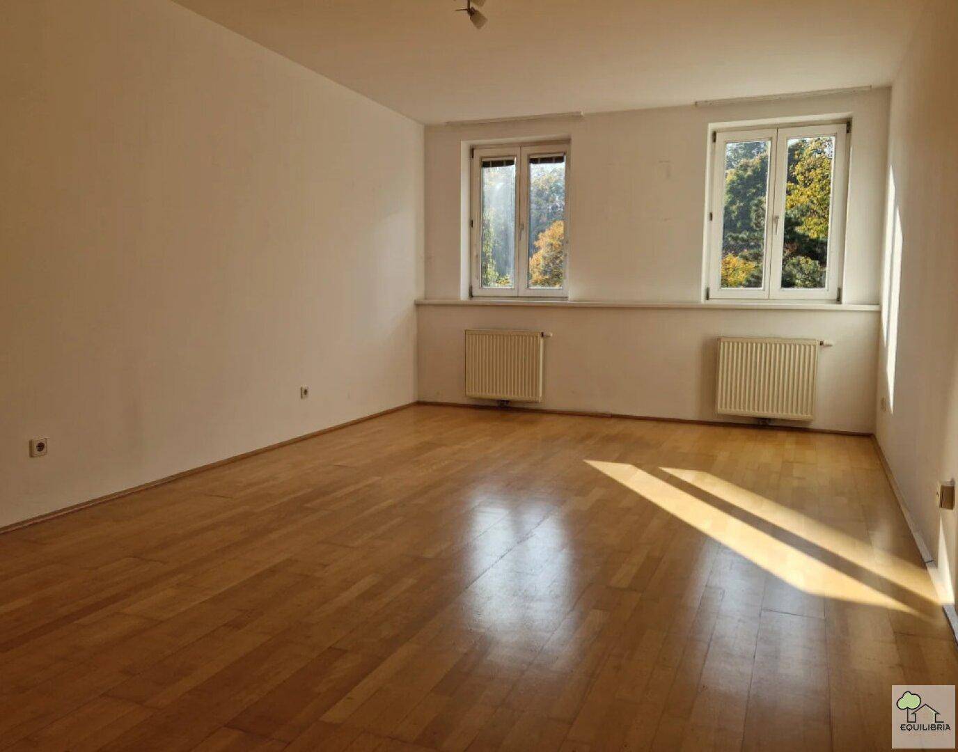 Geräumiges Wohnzimmer mit hellem Parkettboden und zwei Fenstern mit Blick ins Grüne.