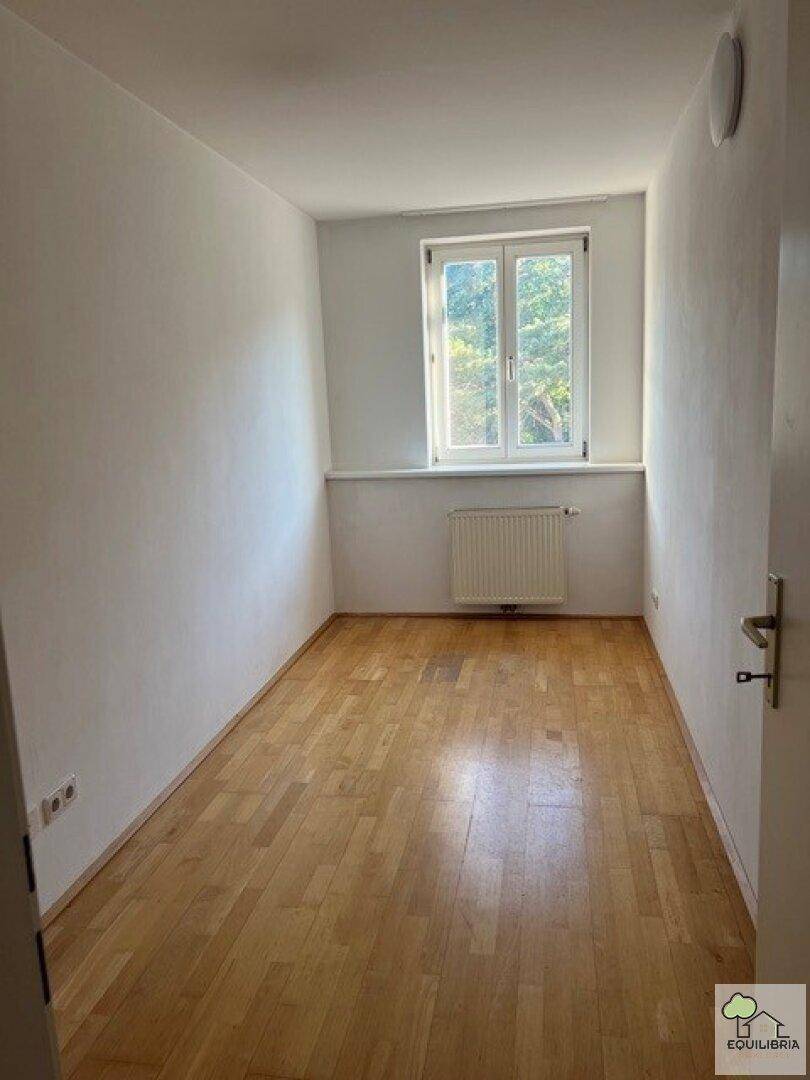 Kleiner, heller Raum mit Parkettboden und einem Fenster mit Blick ins Grüne.