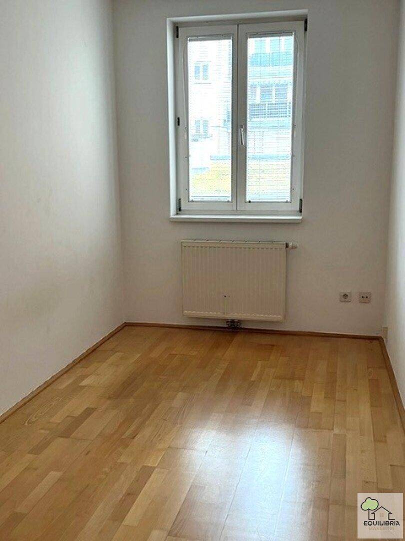 Helles Zimmer mit Parkettboden und Fenster, das einen Blick auf gegenüberliegende Gebäude bietet.