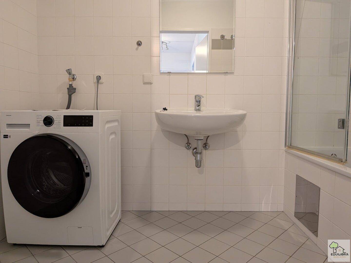 Badezimmer mit weißen Fliesen, Waschbecken, Spiegel und Waschmaschine, sowie Duschbereich.