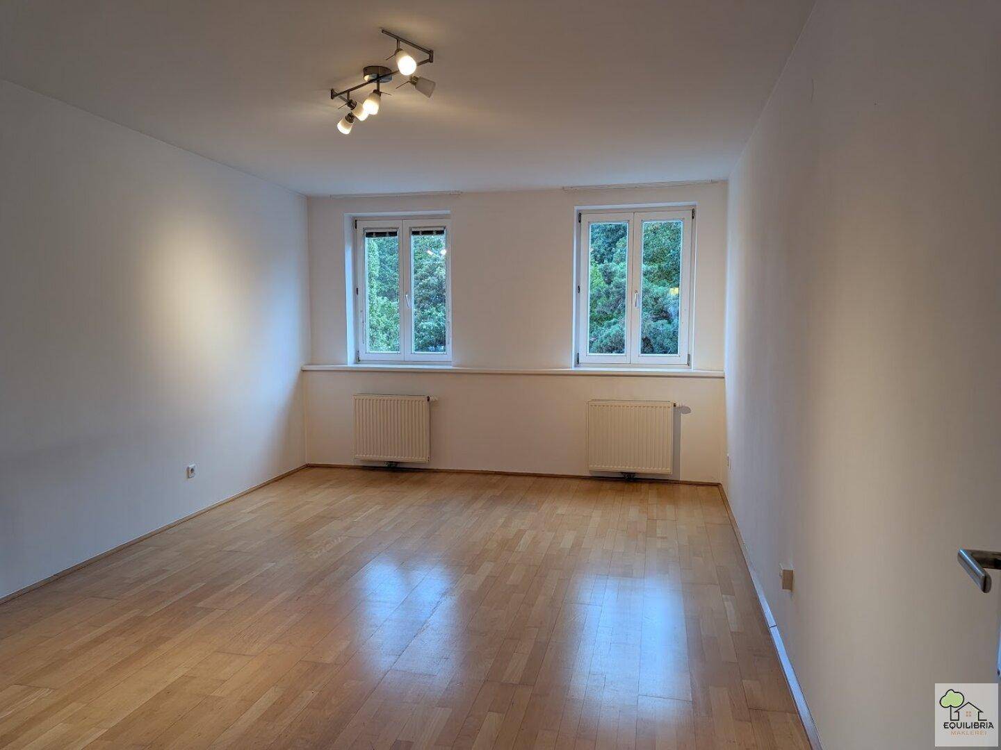 Helles Zimmer mit Parkettboden, weißen Wänden und zwei Fenstern mit Blick ins Grüne.
