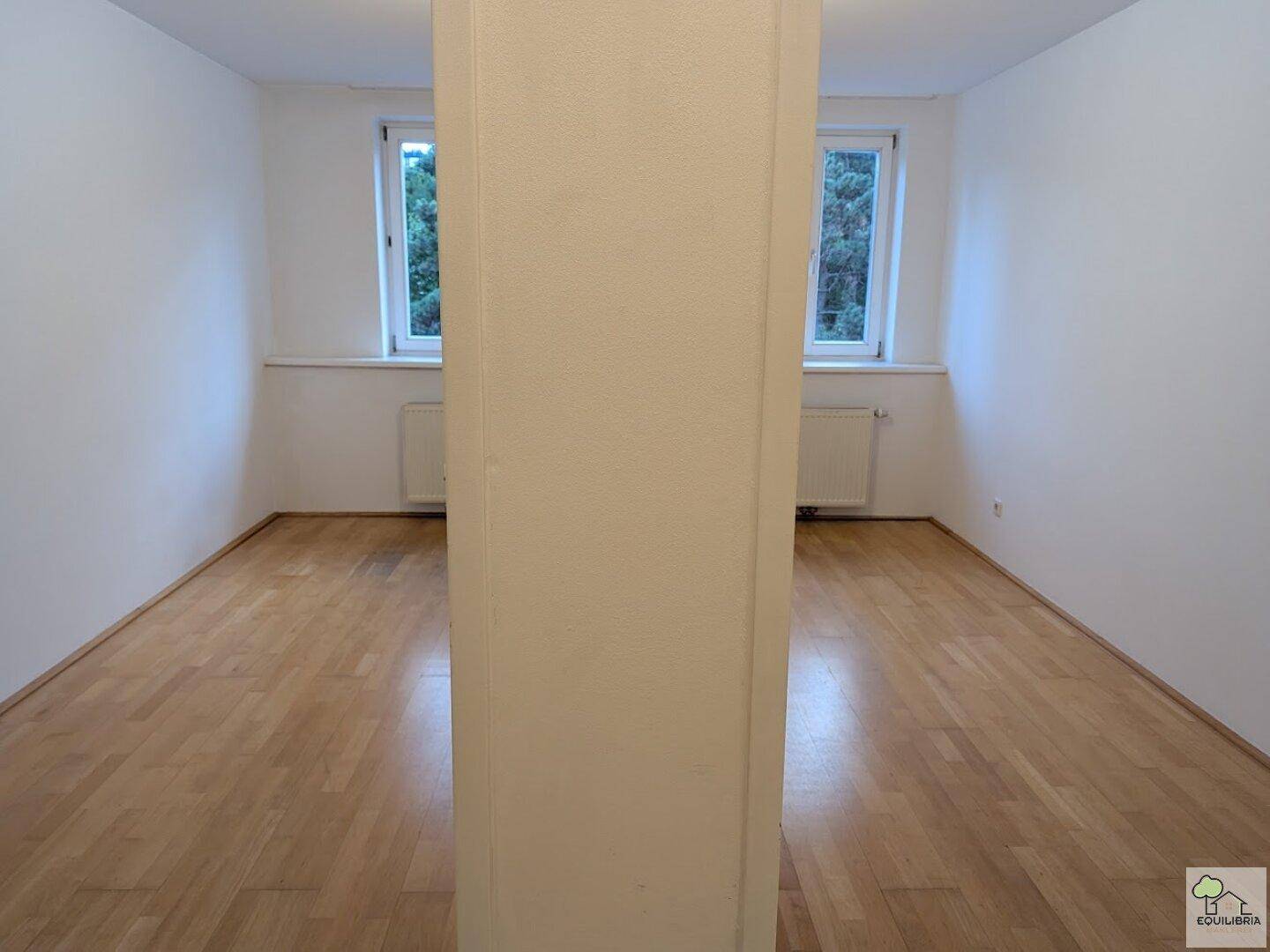 Heller Raum mit Parkettboden, weißen Wänden und Fenstern mit Blick auf Bäume.