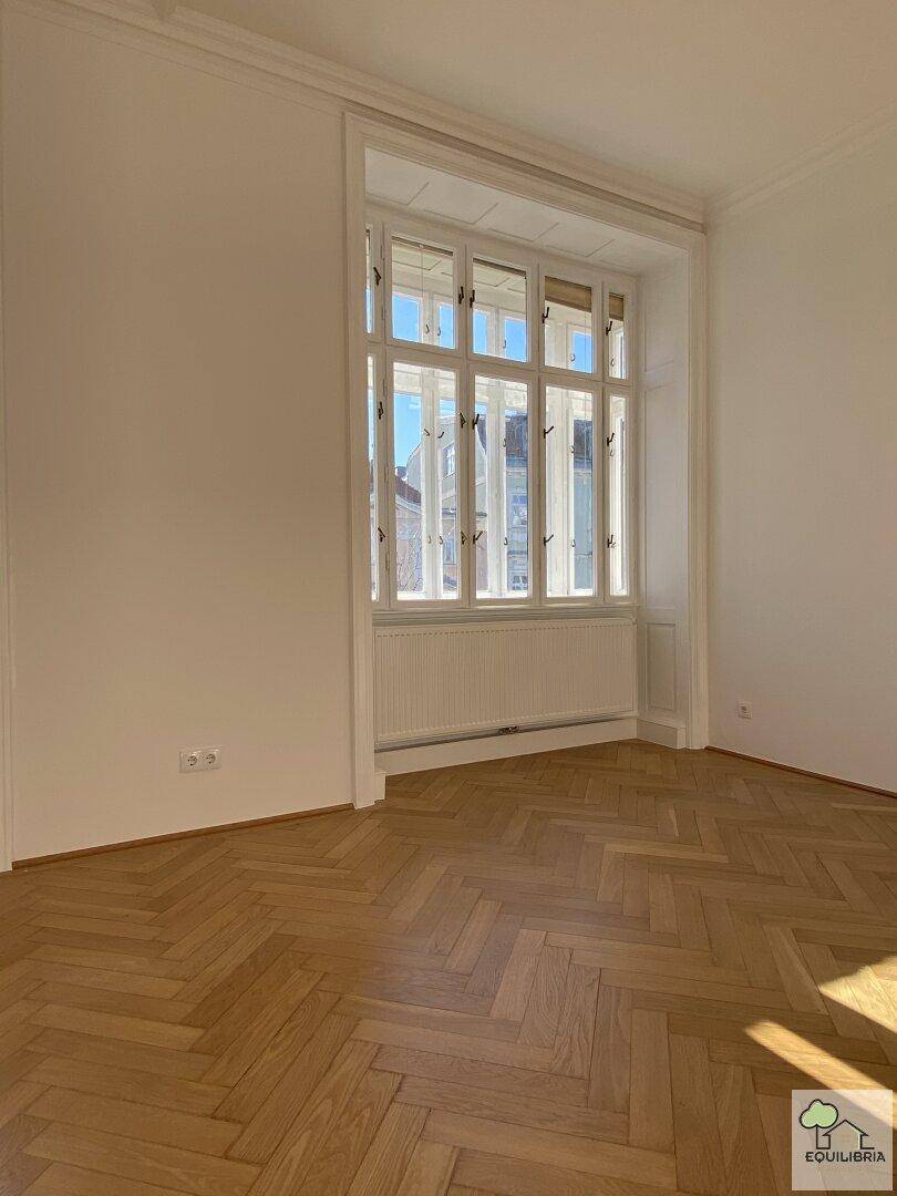 Helles Zimmer mit Fischgrätparkett und einem großen, mehrteiligen Fenster.