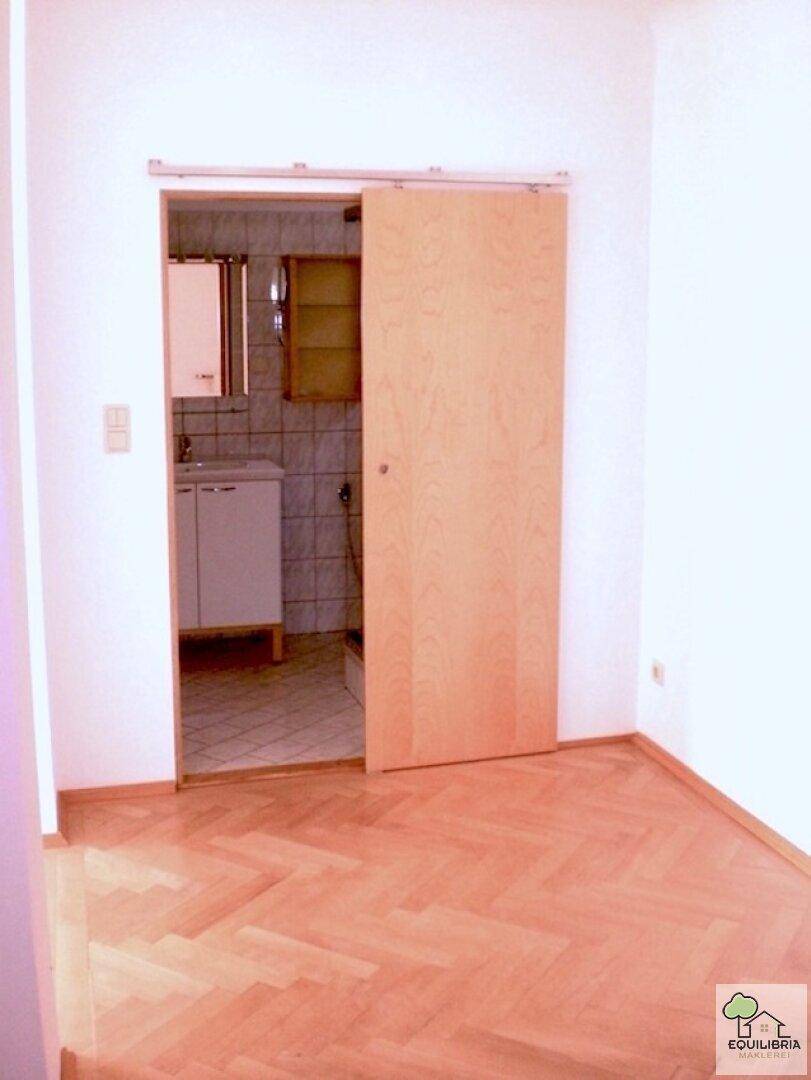 Raum mit Parkettboden und Schiebetür, die zu einem gefliesten Badezimmer führt.