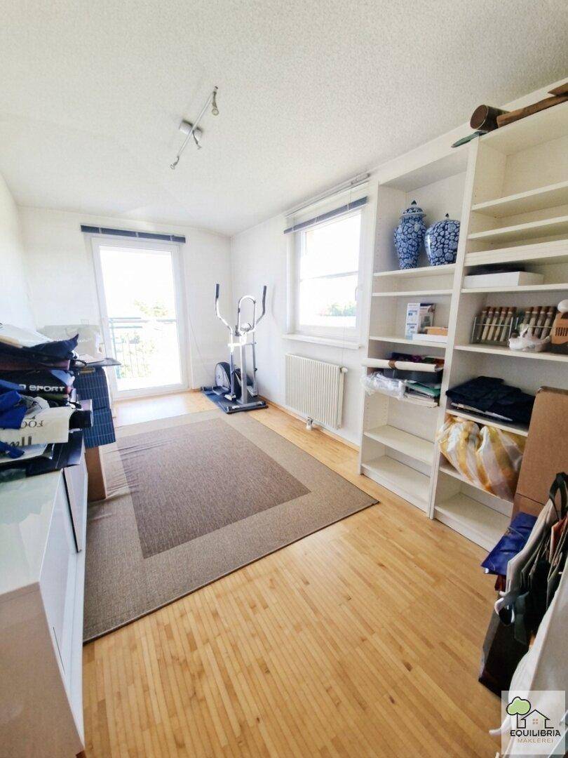Helles Zimmer mit Regalsystem, Sportgerät und Zugang zu einem kleinen Balkon.