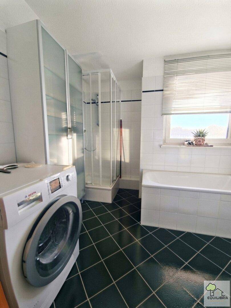 Geräumiges Badezimmer mit dunklen Fliesen, Badewanne, Duschkabine und Waschmaschine.