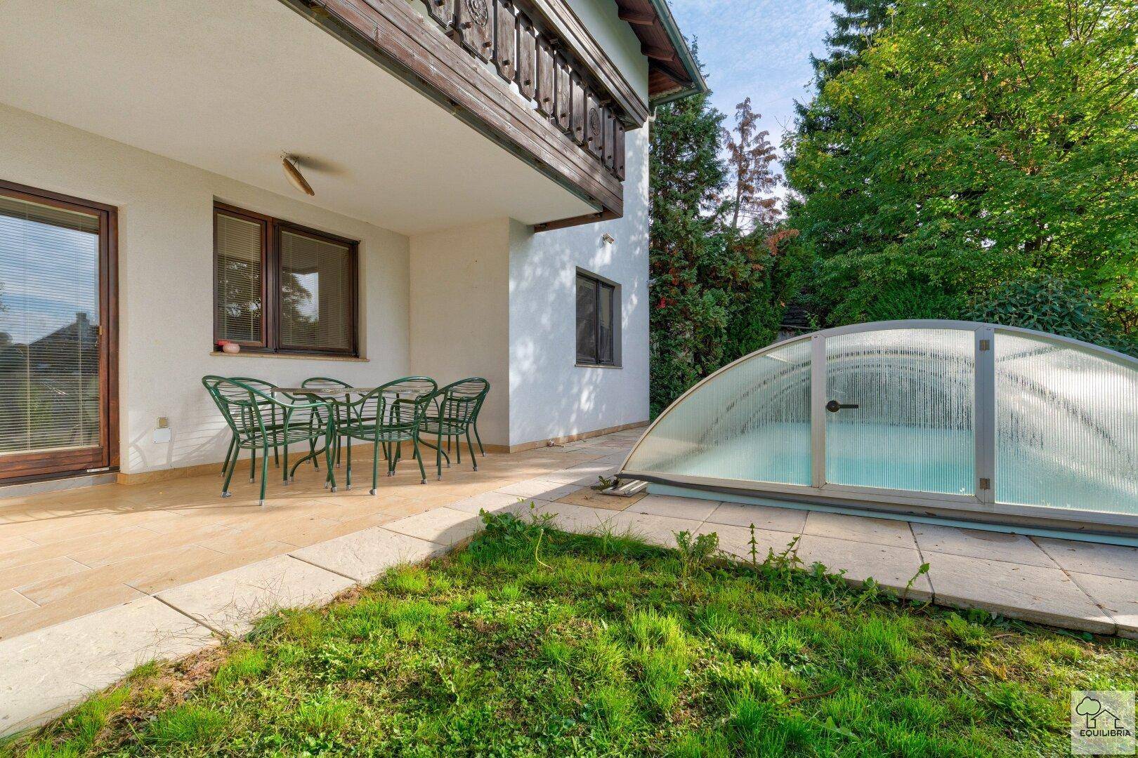 Sonniger Terrassenbereich mit Sitzgelegenheiten und einem überdachten Schwimmbecken im Garten.