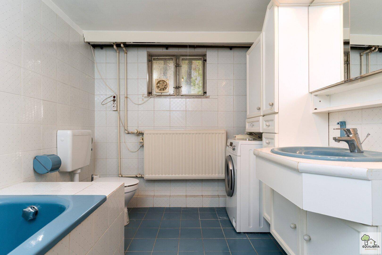 Funktionales Badezimmer im 80er-Jahre-Stil mit blauer Badewanne, WC und Waschmaschine.