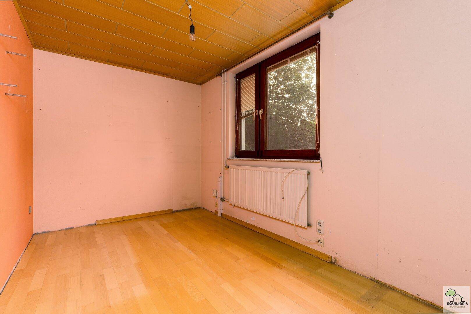 Einfaches Zimmer mit Holzdecke, Parkettboden und Fenster, das auf den Garten blickt.