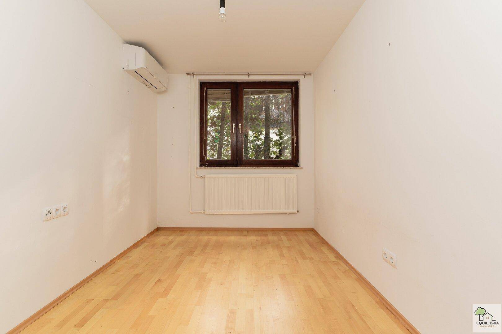 Helles Zimmer mit Parkettboden, Klimaanlage und Blick ins Grüne durch das Fenster.