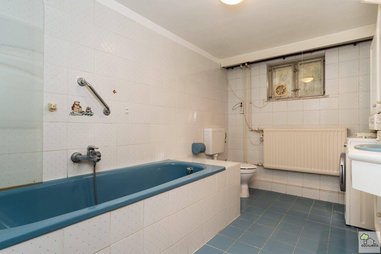 Badezimmer mit blauer Badewanne, weißen Fliesen und Haltegriffen an der Wand.