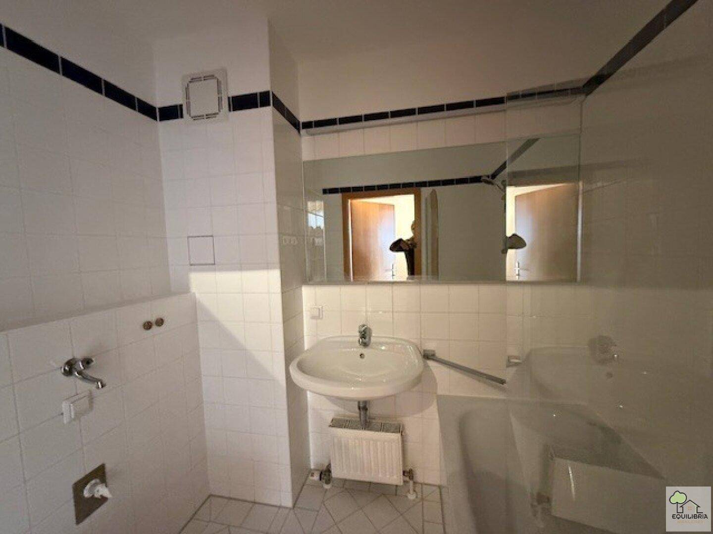 Badezimmer mit Waschbecken, großem Spiegel und Abluftventilator, in hellen Fliesen gehalten.