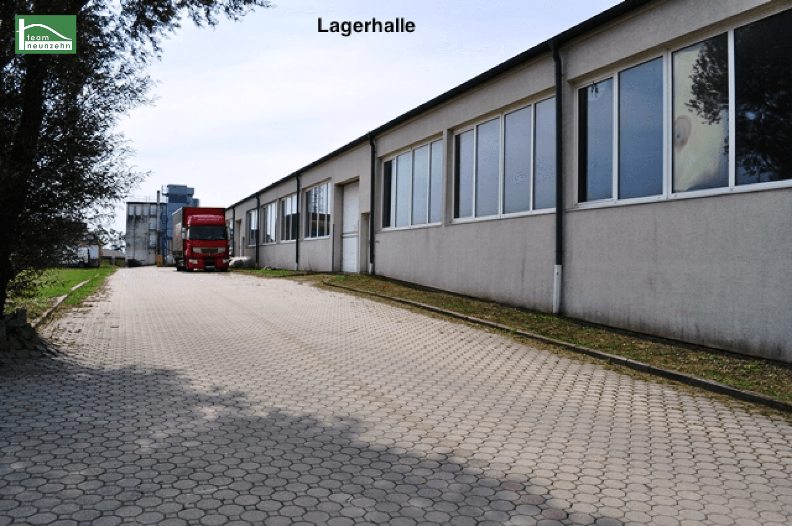 Lange Lagerhalle mit vielen Fenstern und einem gepflasterten Zufahrtsweg, ein LKW ist sichtbar.