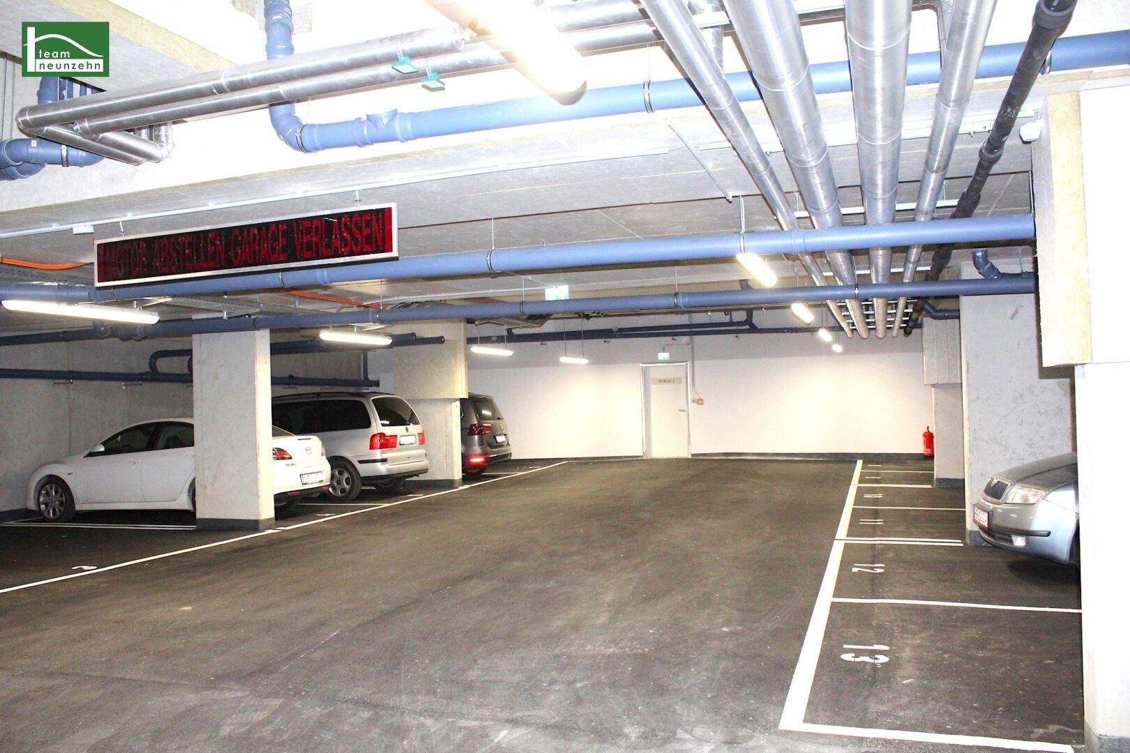 Tiefgarage mit mehreren Stellplätzen, sichtbaren Leitungen und Beleuchtung.