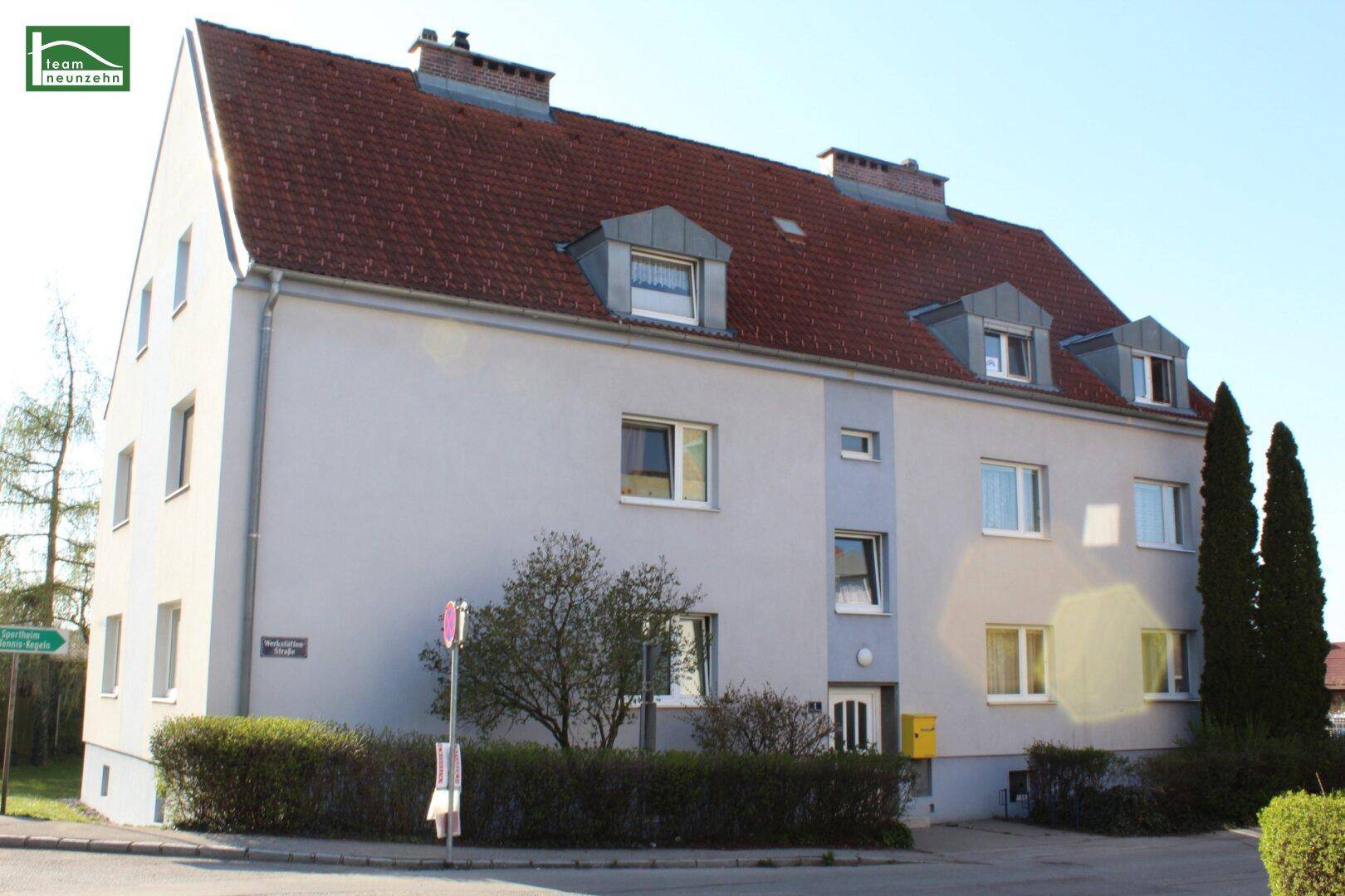 Seitenansicht des Wohngebäudes mit rotem Ziegeldach und hellgrauer Fassade unter blauem Himmel.