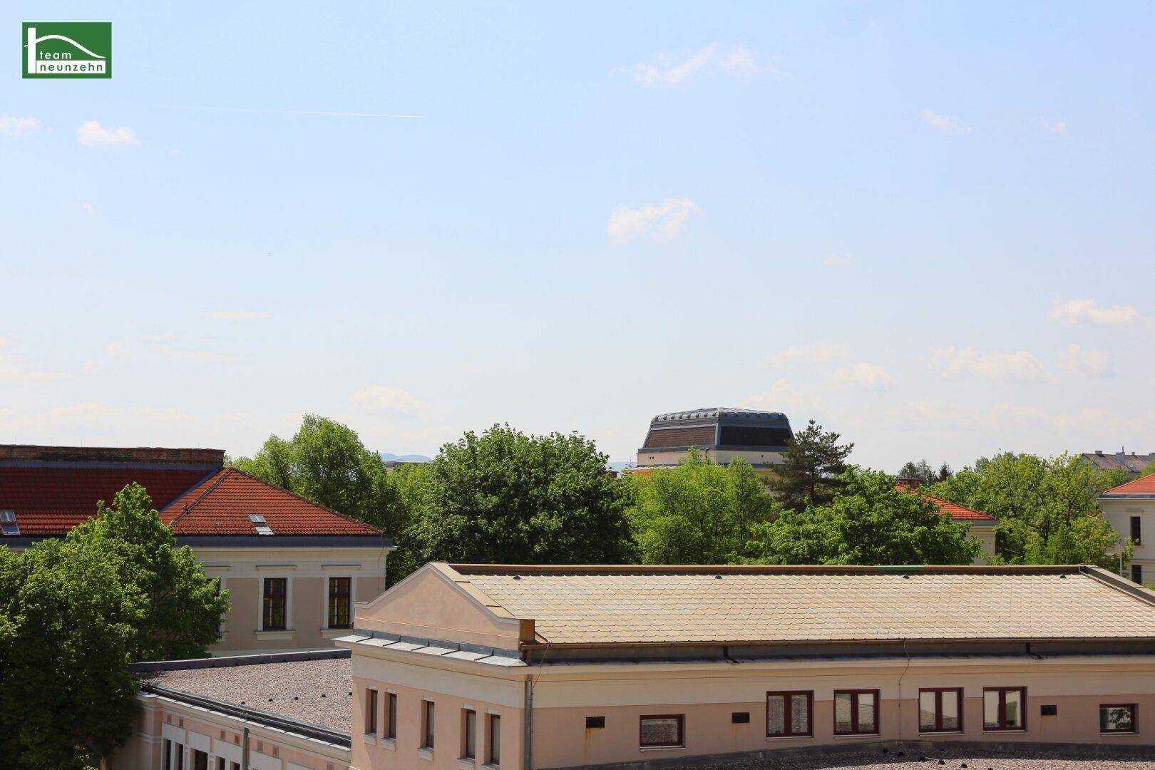 Weitblick über Dächer und grüne Baumwipfel unter einem klaren blauen Himmel.