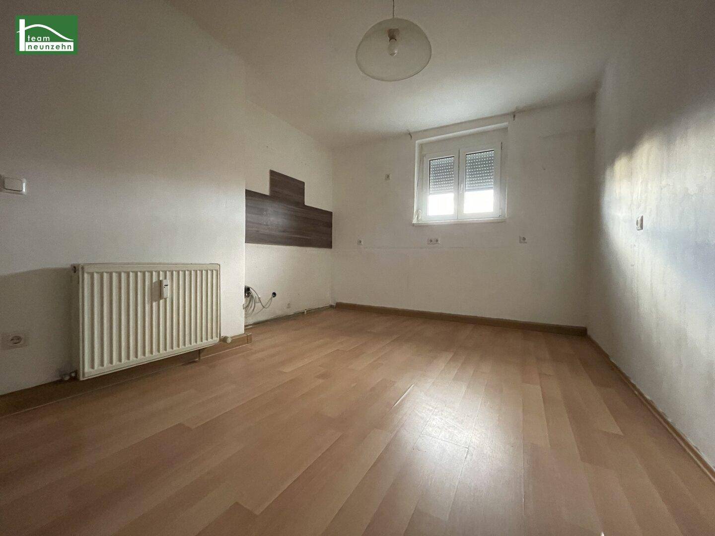 Leerer Raum mit Laminatboden, einem Fenster mit Rollläden und einem Heizkörper an der Wand.