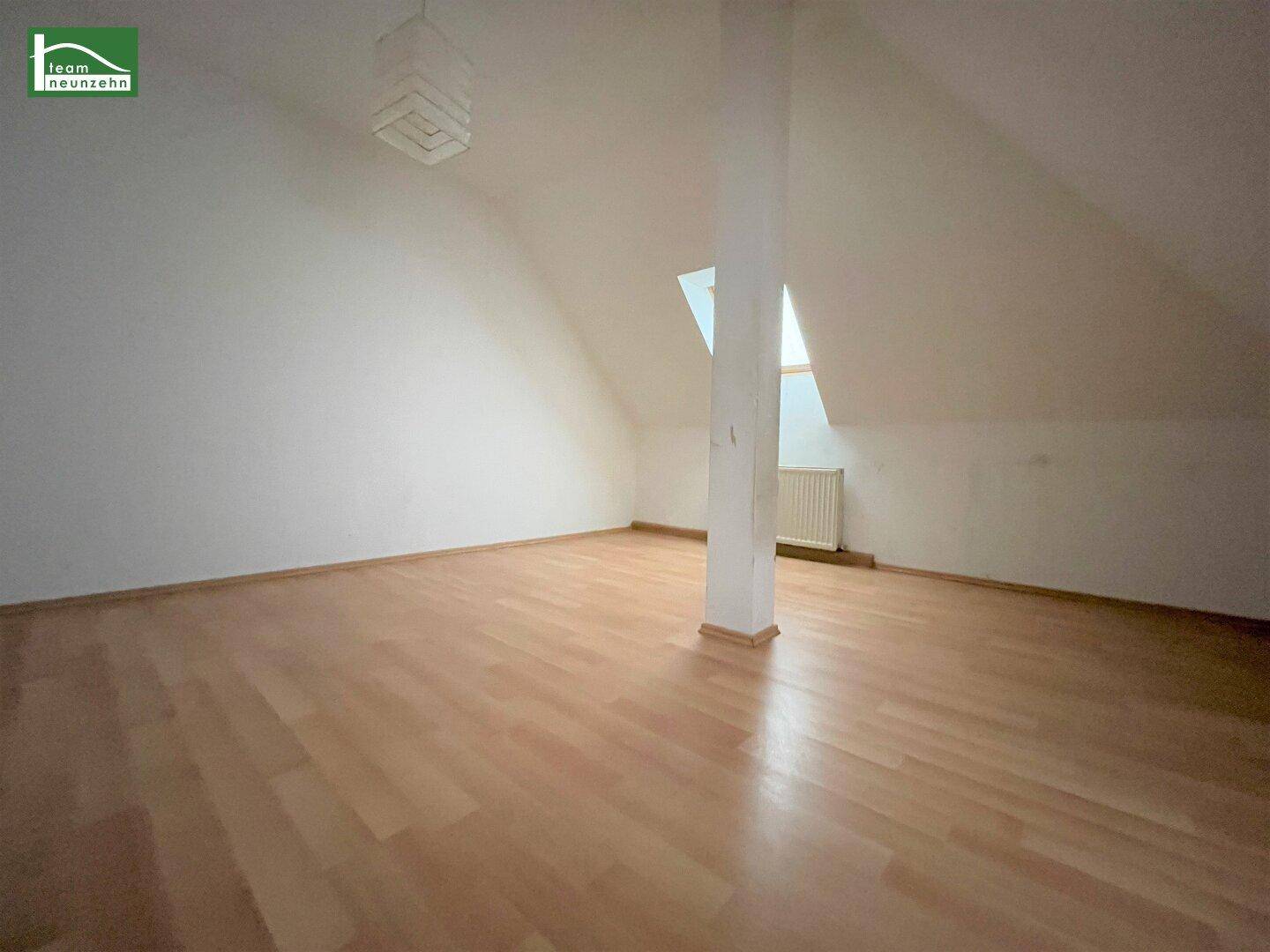 Geräumiger Dachgeschossraum mit Laminatboden, Dachschrägen und einem Heizkörper unter dem Fenster.