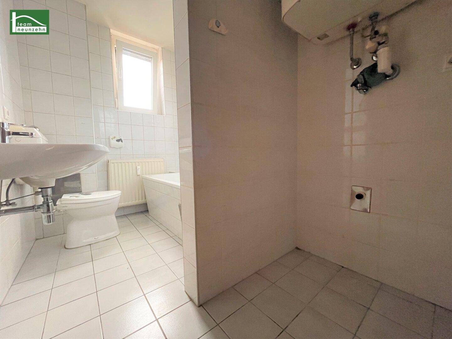 Helles Badezimmer mit weißen Fliesen, Waschbecken, Toilette und einem Fenster, teilweise durch eine Wand getrennt.