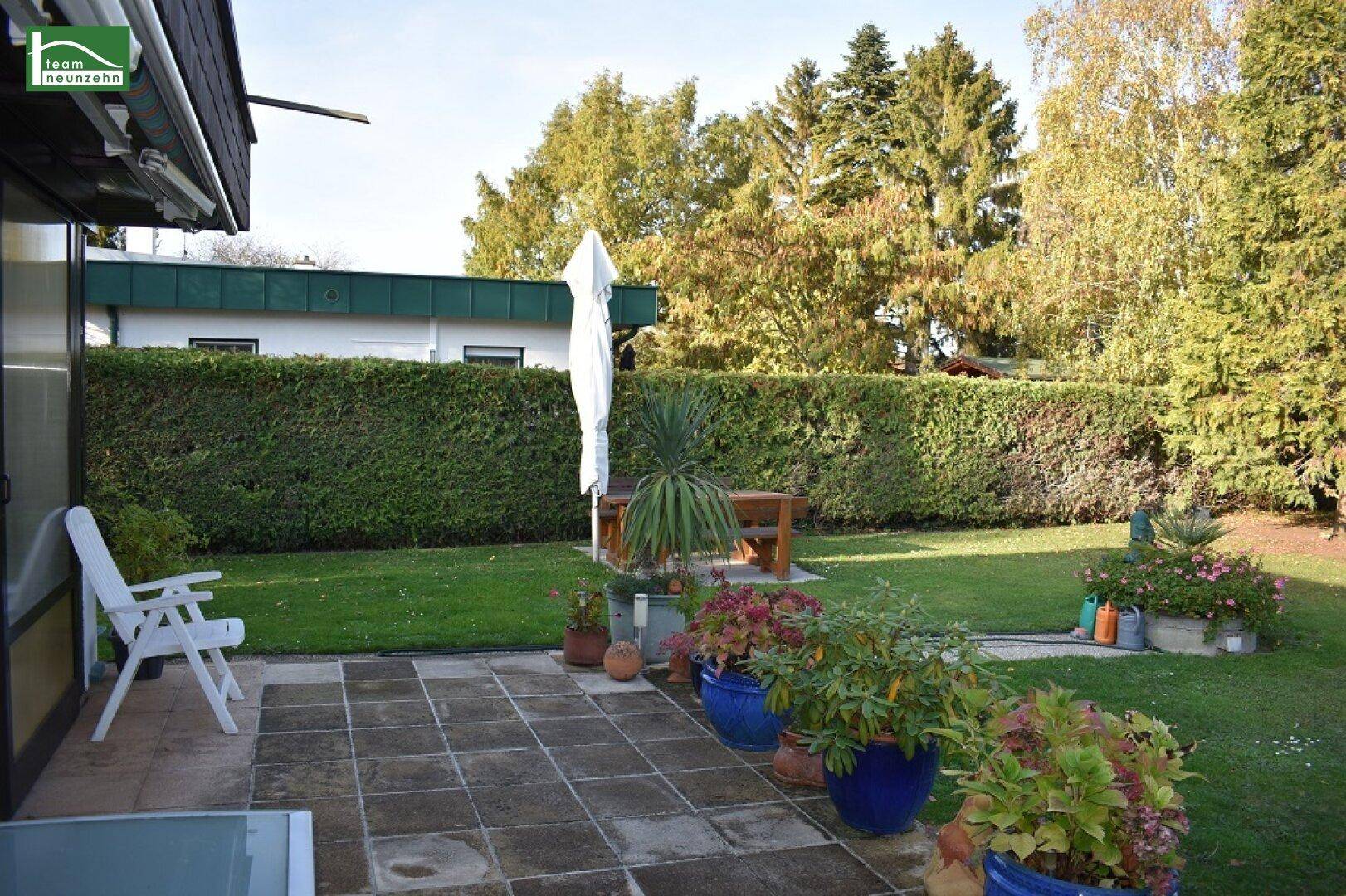 Gedeckte Terrasse mit Fliesenboden, vielen Topfpflanzen und Blick auf den grünen Gartenbereich.