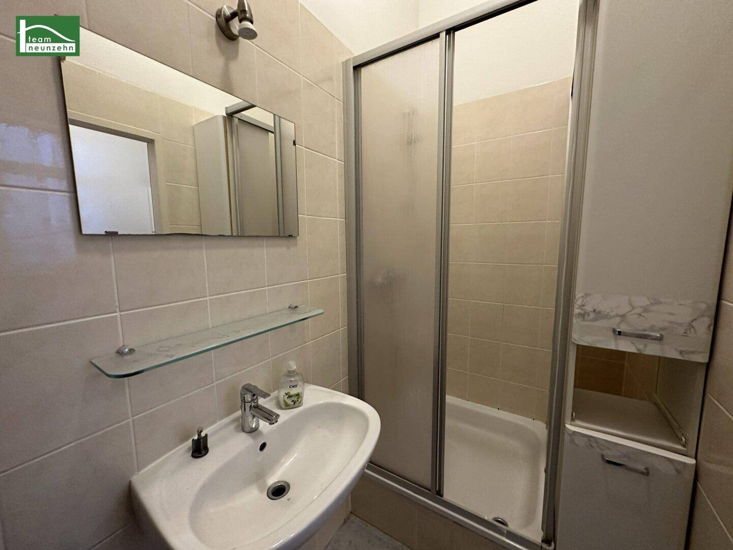 Badezimmer mit beigefarbenen Fliesen, Waschbecken, Spiegel und einer praktischen Duschkabine.