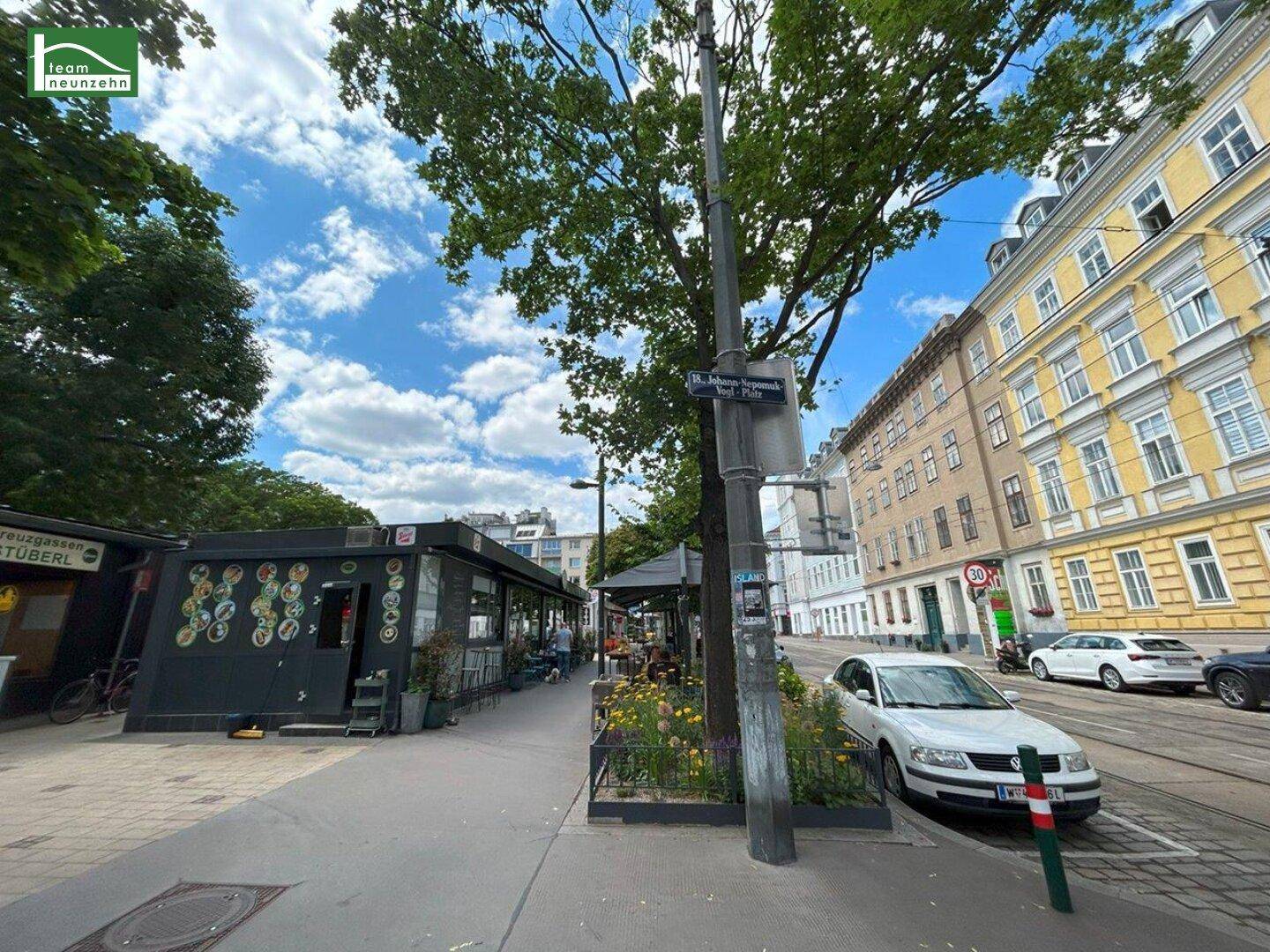 Straßenansicht mit historischen Gebäuden, parkenden Autos und einem Straßenschild unter Bäumen.