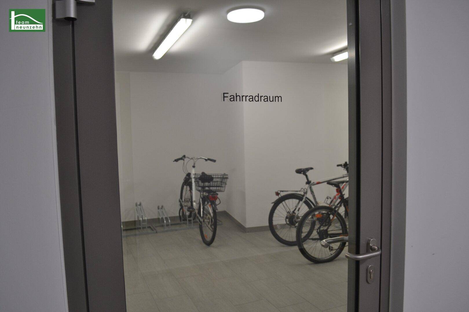 Heller Fahrradraum mit mehreren Fahrrädern und Fliesenboden, durch eine Glastür sichtbar.