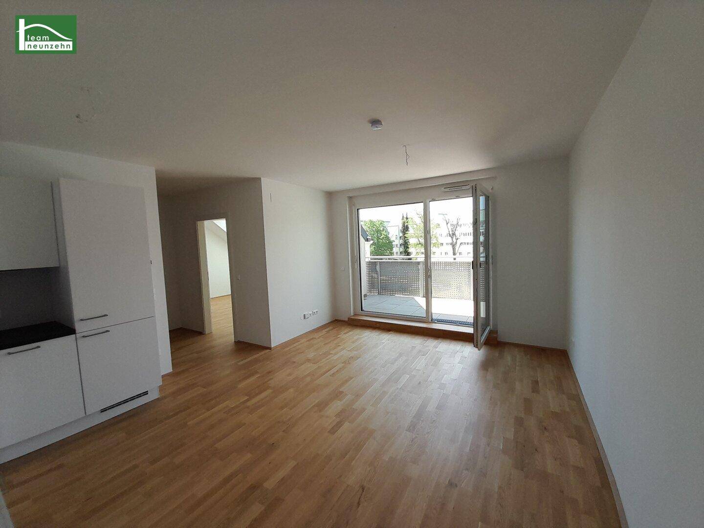 Helles, leeres Wohnzimmer mit Holzboden und großen Schiebetüren zum Balkon.