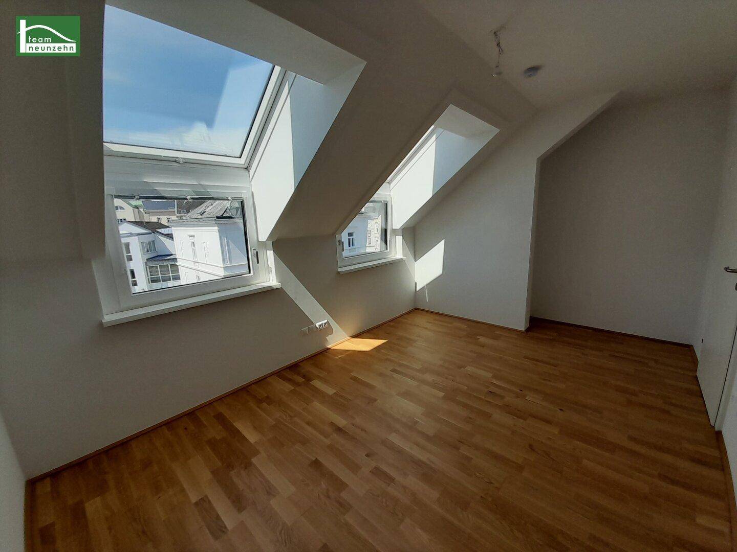 Lichtdurchflutetes Dachzimmer mit Holzboden, schrägen Wänden und Blick auf die Umgebung.