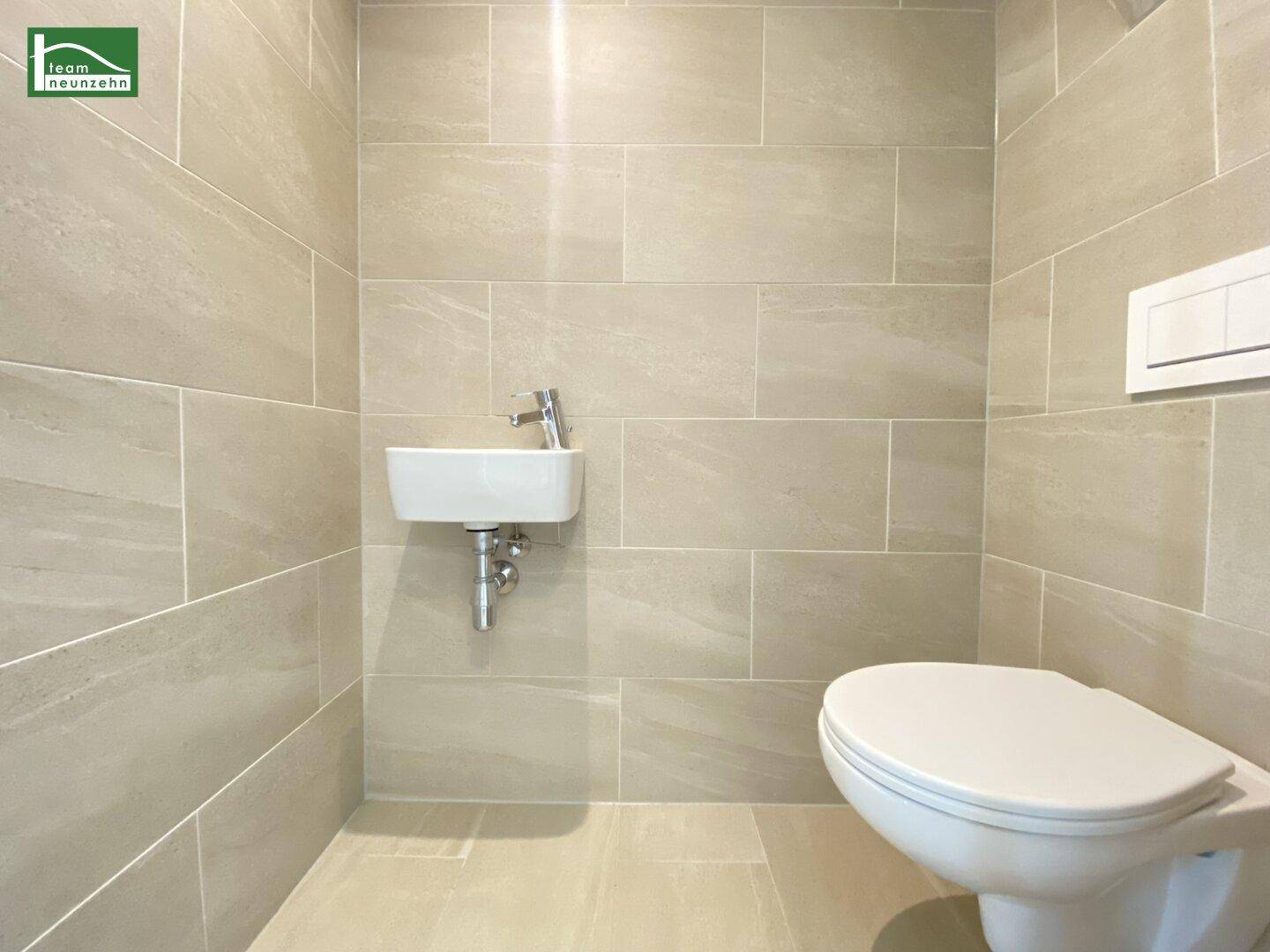 Separates WC mit hellen Fliesen, wandhängender Toilette und kleinem Handwaschbecken.
