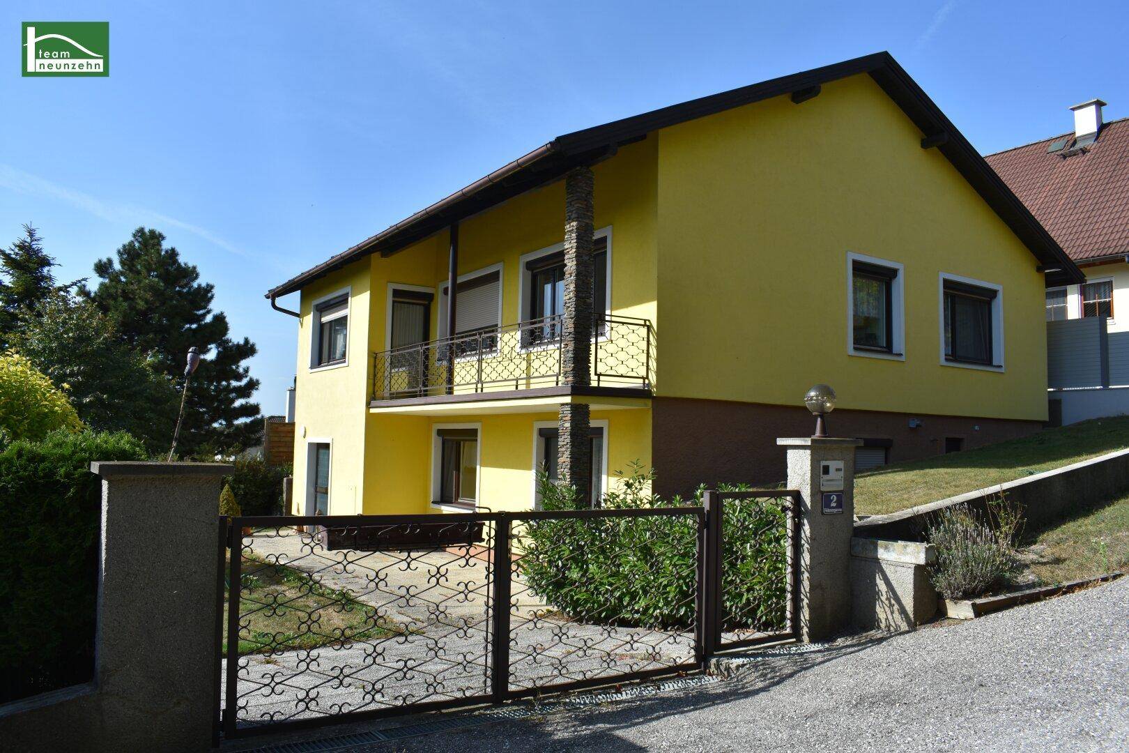 Gelbes Einfamilienhaus mit Balkon, schmiedeeisernem Tor und gepflegtem Vorgarten.