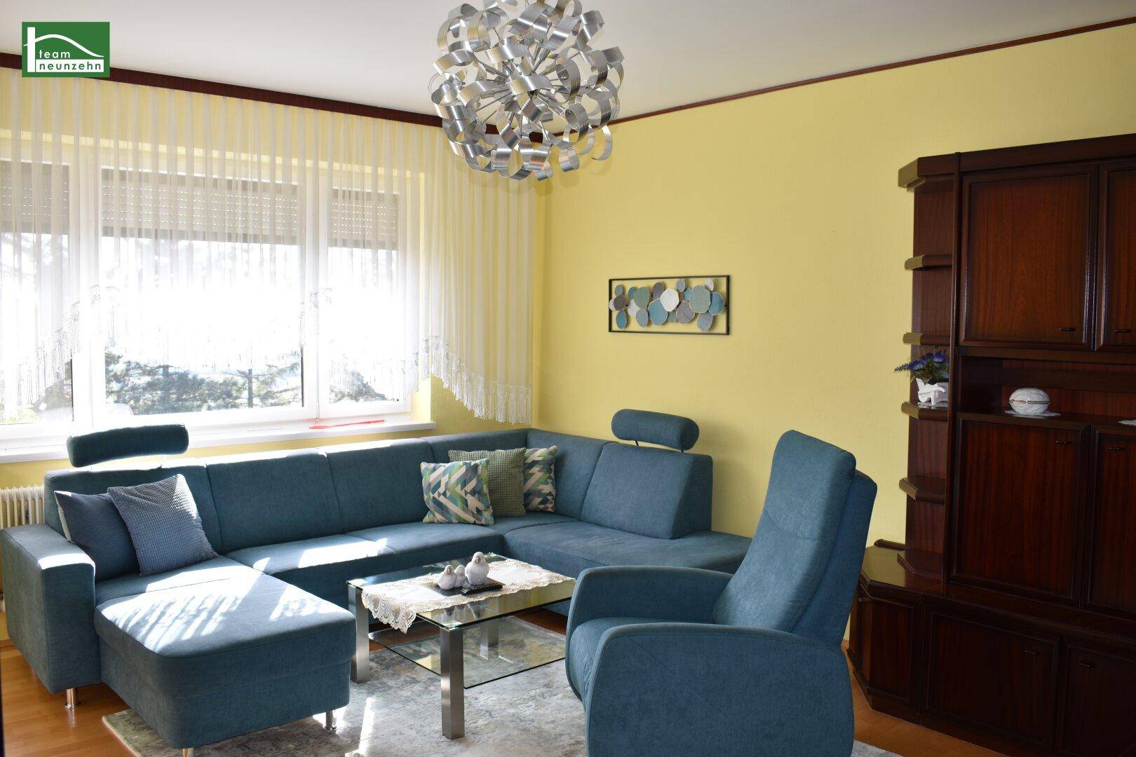 Gemütliches Wohnzimmer mit großem blauen Ecksofa, Glascouchtisch und viel Tageslicht.