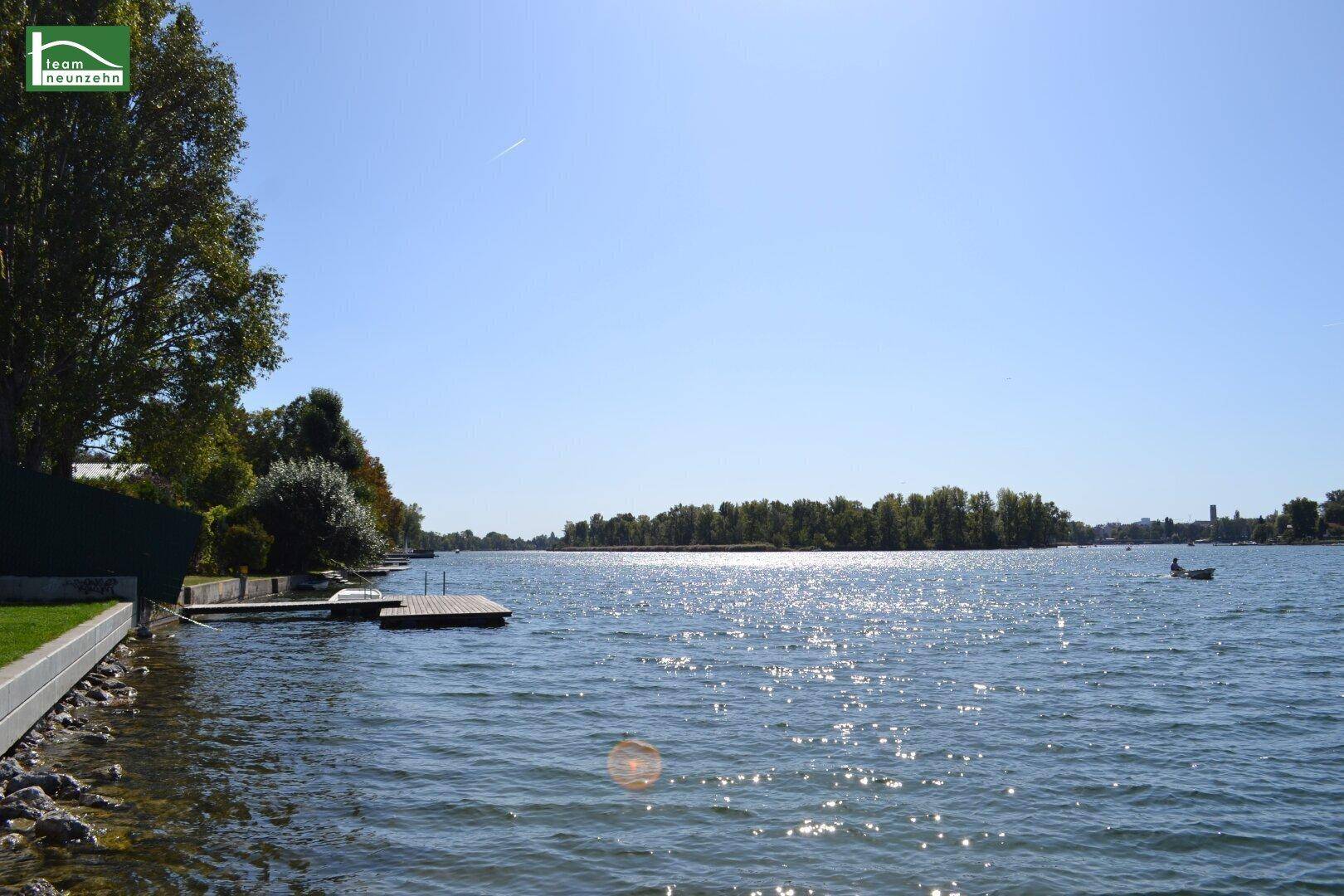 Weitläufiger Blick auf das glitzernde Wasser mit Steg und grünen Ufern unter blauem Himmel.