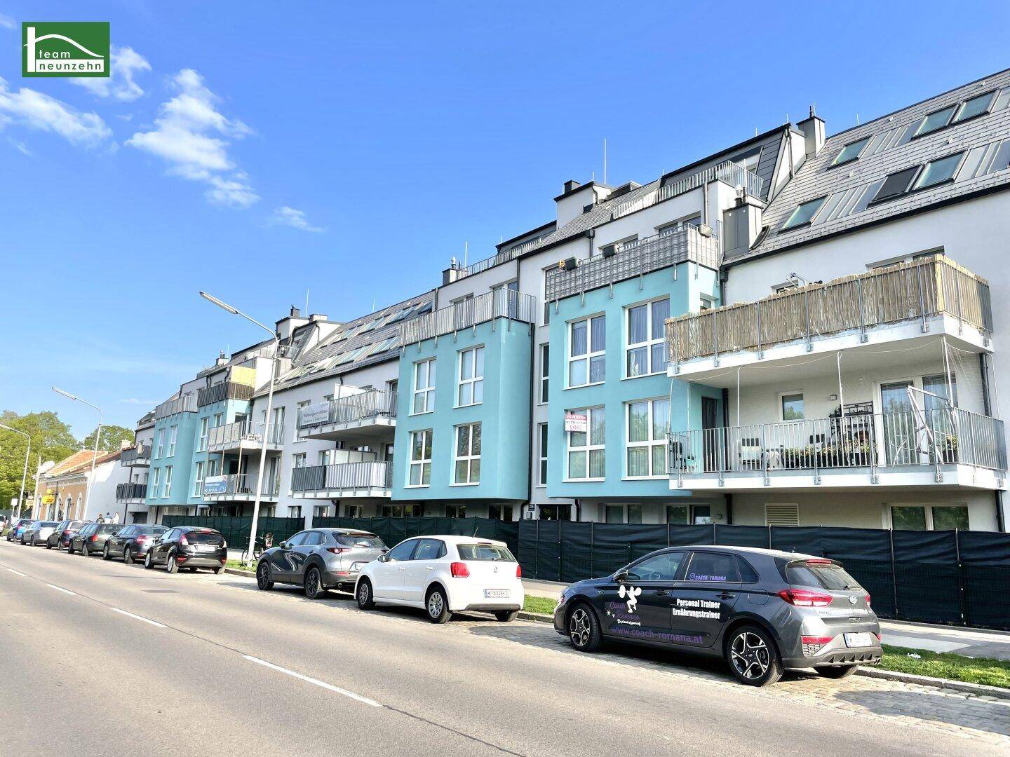 Moderne Wohngebäude-Fassade mit blauen und weißen Akzenten, Balkonen und parkenden Autos entlang der Straße.