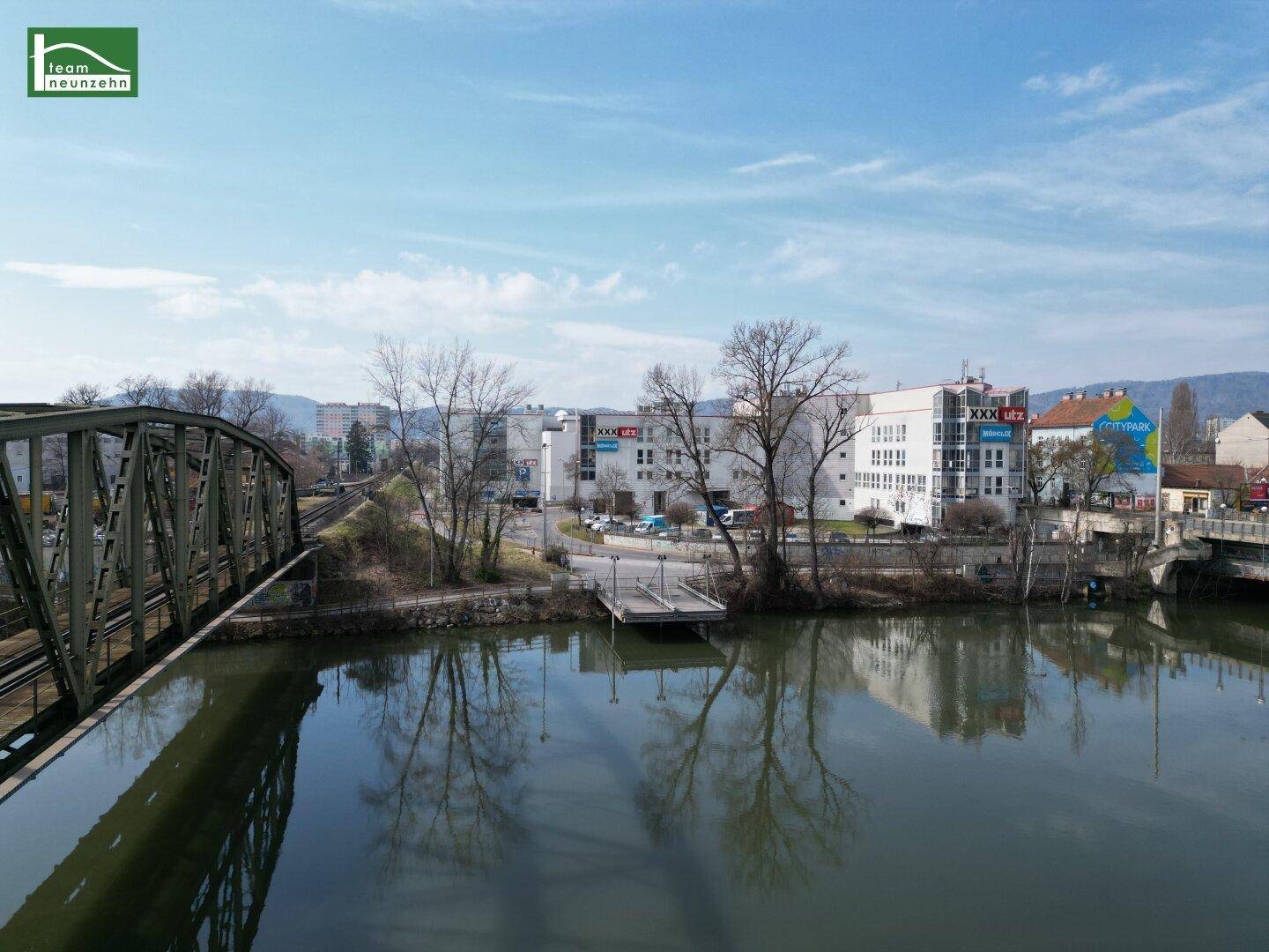 Blick über einen Fluss mit einer Brücke und städtischer Bebauung im Hintergrund unter blauem Himmel.