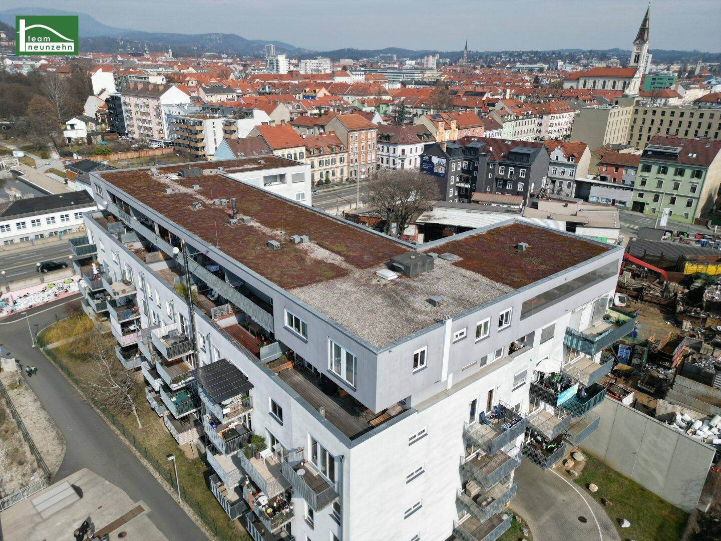 Luftaufnahme eines modernen Wohngebäudes mit begrüntem Flachdach und Stadtpanorama im Hintergrund.