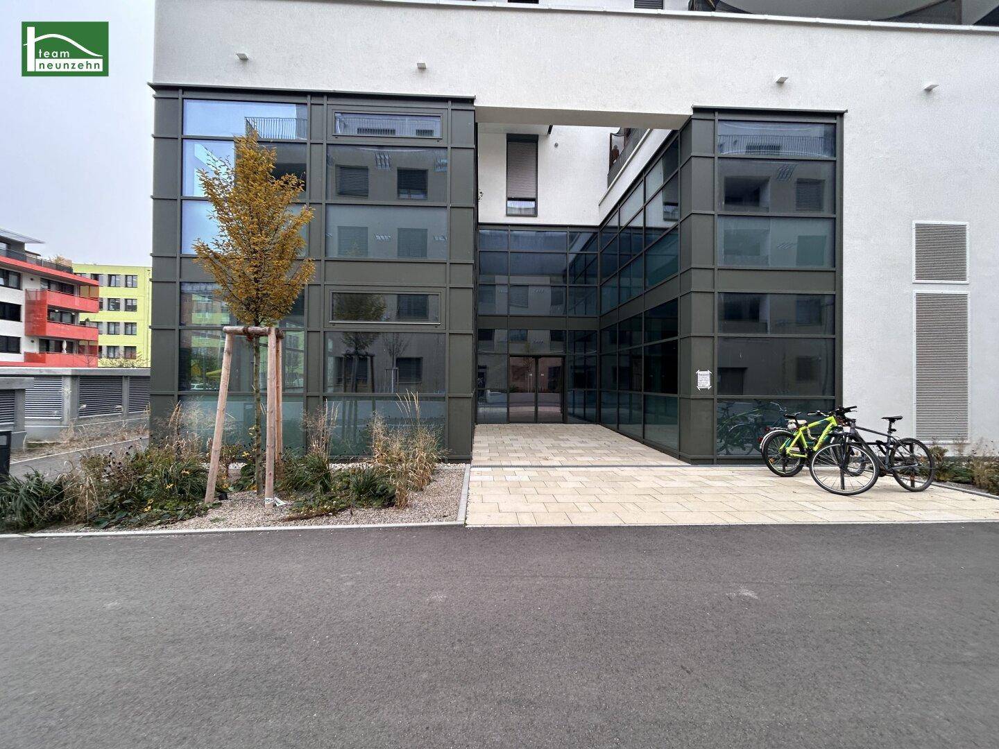 Moderner Eingangsbereich eines Gebäudes mit viel Glas und klaren Linien, umgeben von junger Bepflanzung.