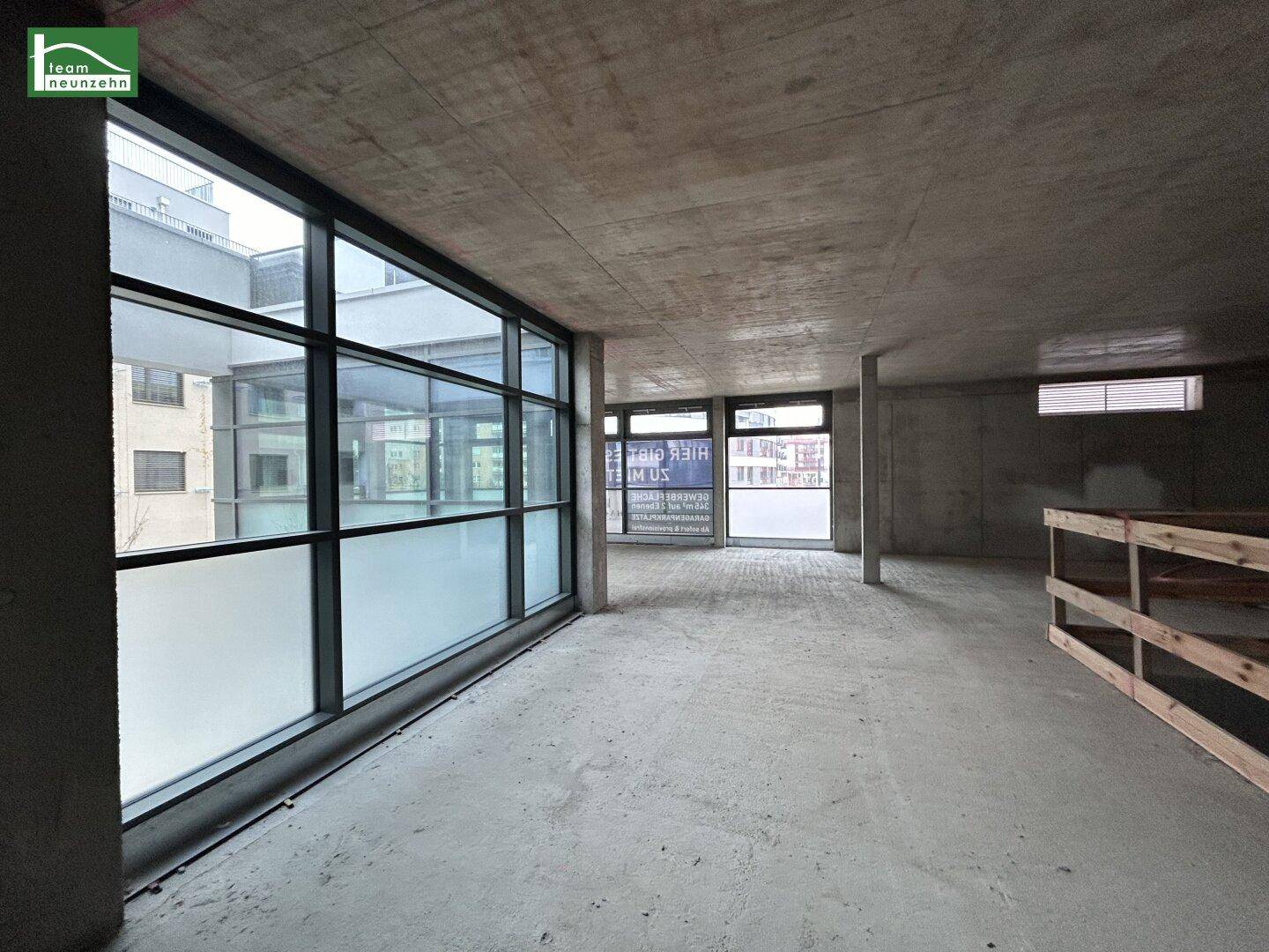Unfertiger Gewerberaum mit Betonboden und -decke, großen Fenstern und Holzregalen im Hintergrund.