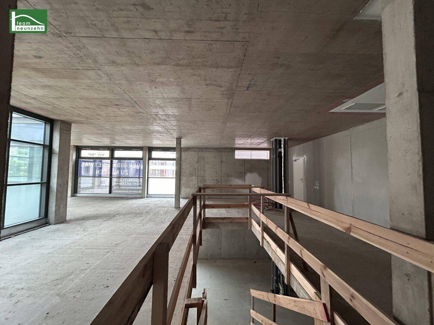 Großer Gewerberaum im Rohbau mit Betonoberflächen, einer Rampe mit Holzgeländer und großen Fenstern.