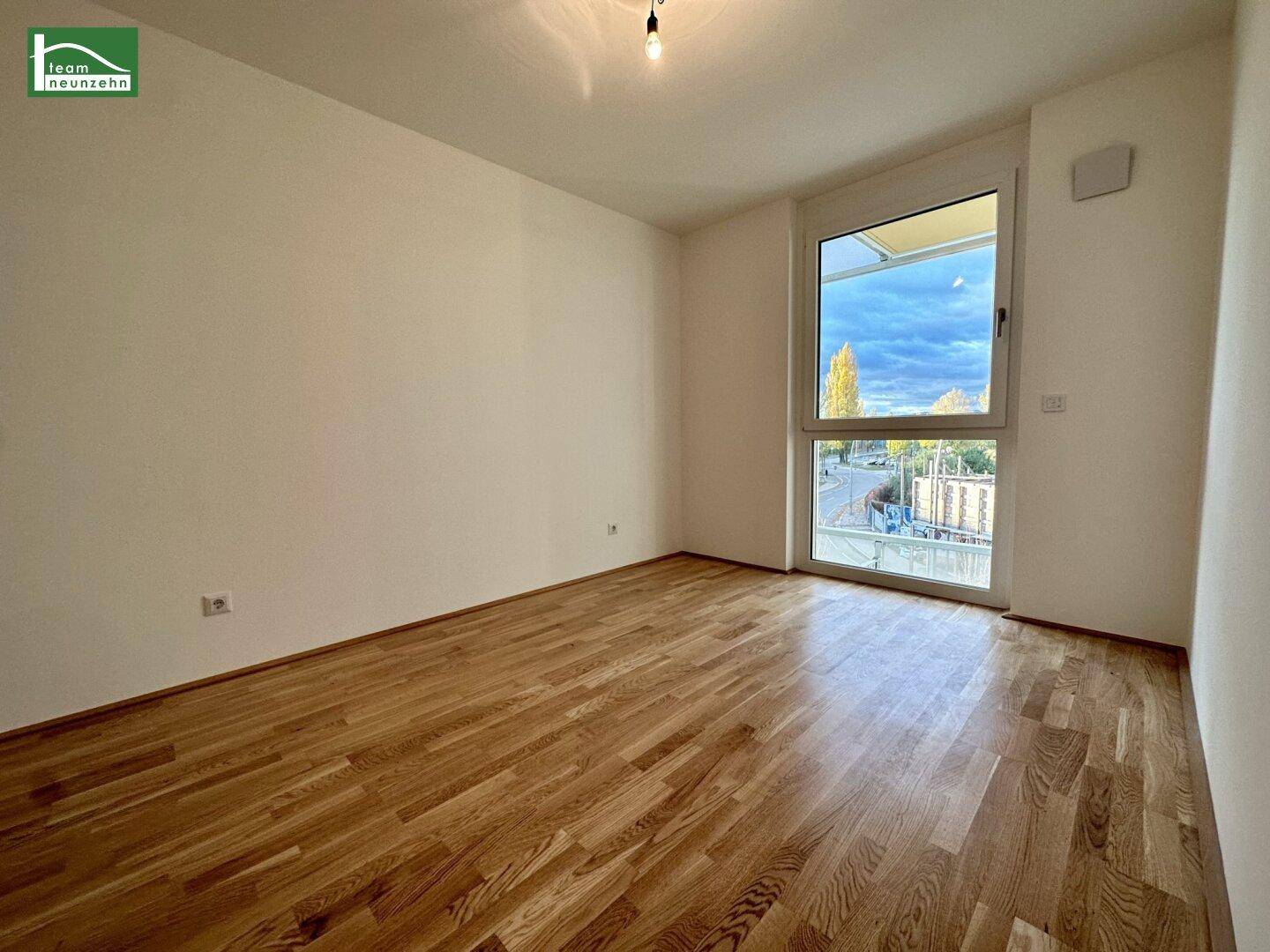 Leeres Zimmer mit Holzboden und großem Fenster, das viel Tageslicht hereinlässt.
