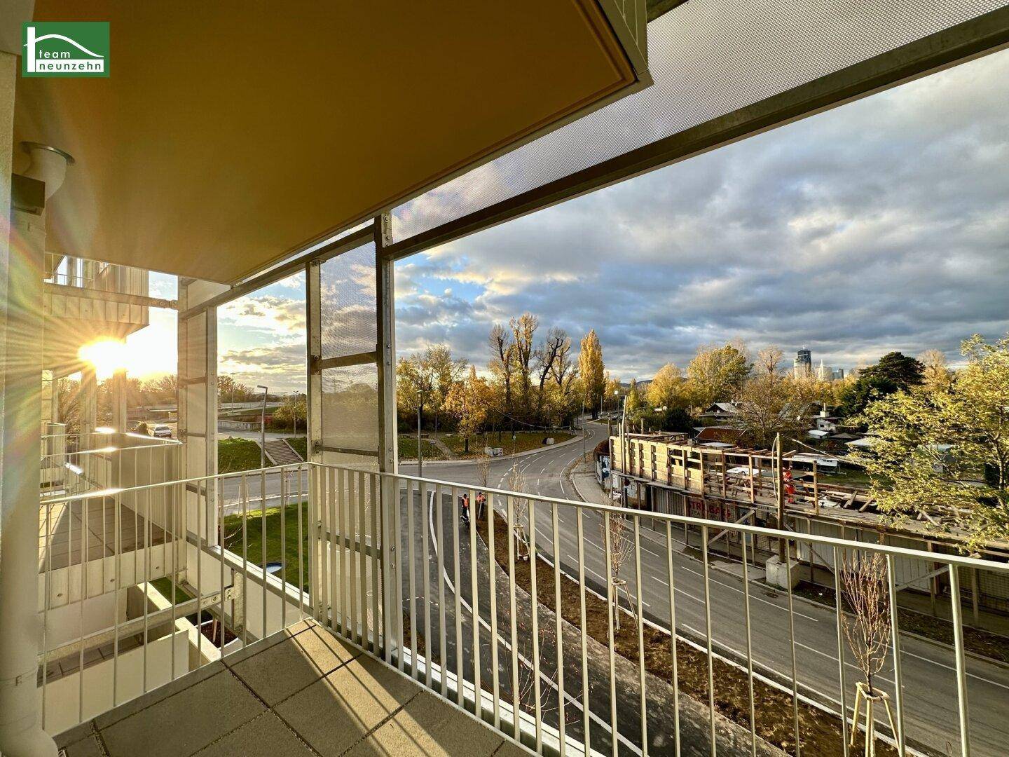 Balkon mit Metallgeländer und Blick auf die Straße und die umliegende Bebauung bei Sonnenuntergang.