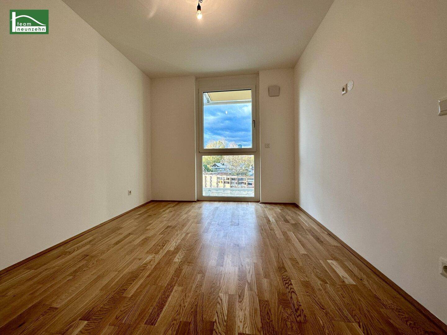 Helles, leeres Zimmer mit Parkettboden und Fenster mit Blick auf die Umgebung.