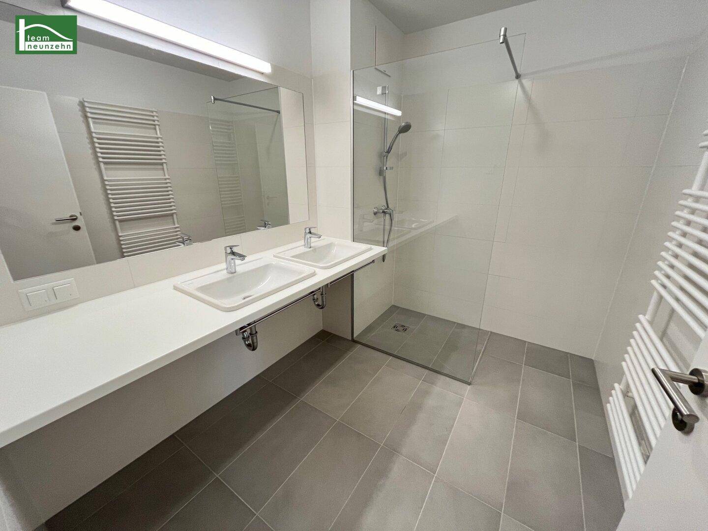Modernes Badezimmer mit Doppelwaschbecken, großer Dusche und Handtuchheizkörper, alles in hellen Tönen gehalten.